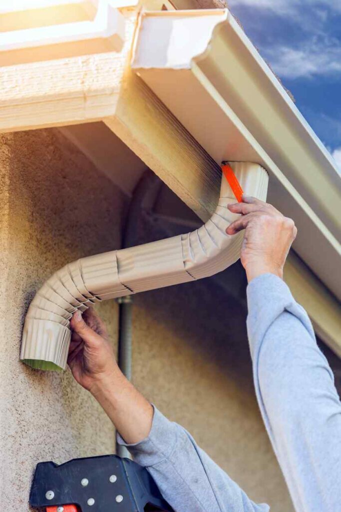 Worker attaching aluminum seamless gutters downspout to fascia of house.
