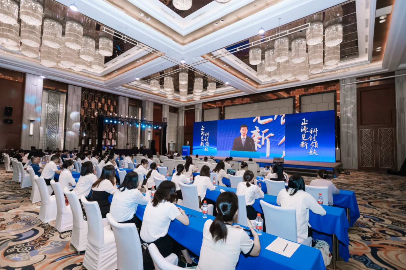 A group of people sitting at tables in a room with a large screenDescription automatically generated with medium confidence