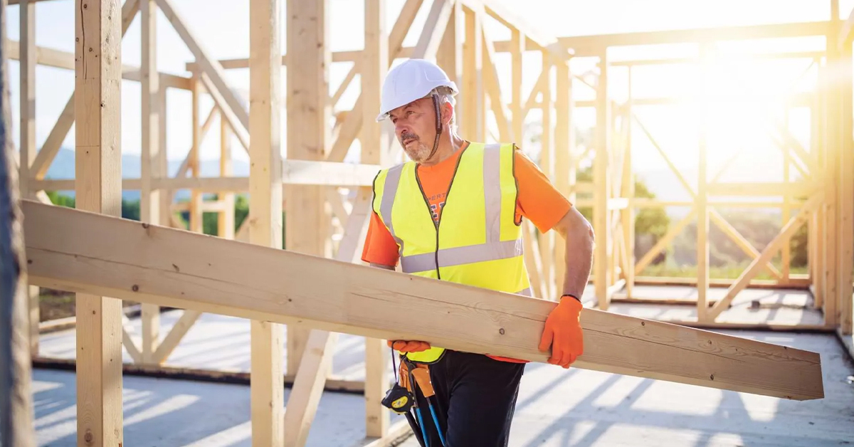 Man wearing construction helmet and high visibility vest carrying wood beam in construction site